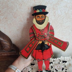 May include: A hand-painted wooden figure of a person wearing a red and gold uniform with a black hat and a black bird on their chest. The figure is standing on a white lace tablecloth.