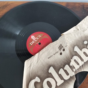 May include: A black vinyl record with a circular red Decca label in the center, partially covered by a torn, beige paper sleeve featuring the word Columbia in large white letters and the text Magic Notes with a musical note symbol.