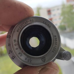 Puede incluir: Un objetivo de c&aacute;mara Industar-50 de color plateado, con ajustes de apertura visibles y el texto "3.5/50" y "ИНДУСТАР-50". El objetivo se sostiene en una mano, con un fondo borroso de una ventana y edificios.