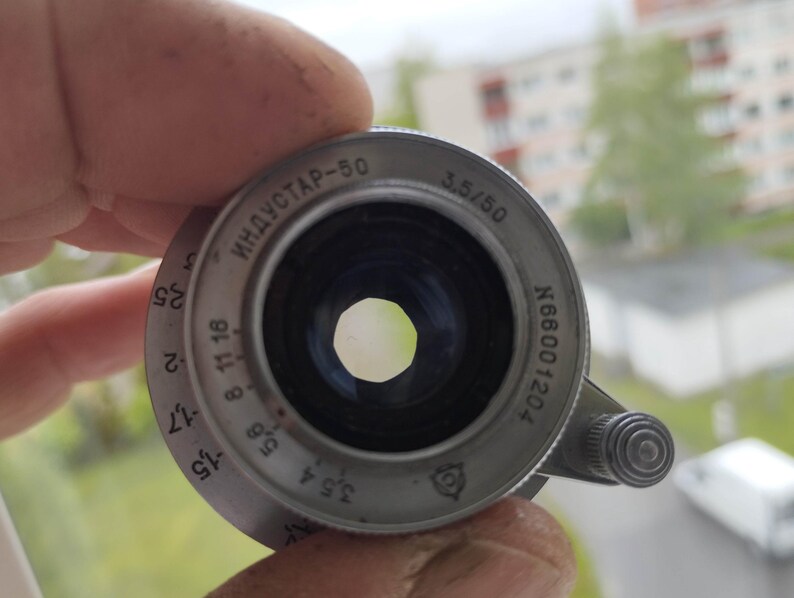 Puede incluir: Un objetivo de c&aacute;mara Industar-50 de color plateado, con ajustes de apertura visibles y el texto "3.5/50" y "ИНДУСТАР-50". El objetivo se sostiene en una mano, con un fondo borroso de una ventana y edificios.