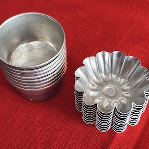 May include: A set of small, round, silver-colored metal baking molds. One stack of molds has a smooth, cylindrical shape, while the other has a fluted, flower-like design. The molds are on a red surface.