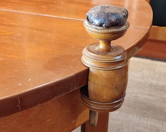 May include: A wooden pin cushion clamped to the edge of a wooden table. The pin cushion is a dark brown, rounded shape, and the clamp is made of turned wood. The table is a warm, medium brown color.