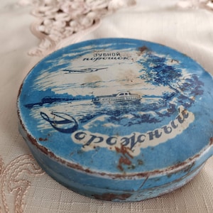 May include: A vintage blue and white tin container with a boat and a plane on the front. The text on the tin reads "зубной порошок" which translates to "tooth powder".