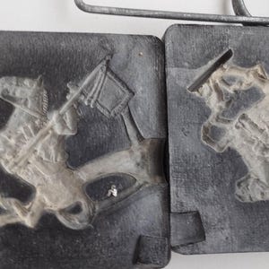 Puede incluir: Un molde de dos piezas para fundir una figurita de metal de una persona a caballo. El molde está hecho de metal gris oscuro y tiene una textura rugosa. El molde está abierto y el interior es visible.