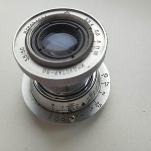Puede incluir: Un objetivo de c&aacute;mara plateado vintage con el texto "ИНДУСТАР-50 3,5/50" y marcas de medici&oacute;n. El objetivo tiene un dise&ntilde;o circular con un elemento de vidrio y un anillo de ajuste de enfoque. El objetivo est&aacute; dise&ntilde;ado para una distancia focal de 50 mm.