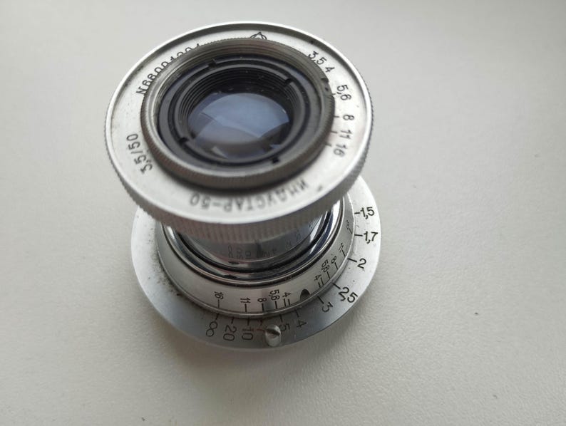 Puede incluir: Un objetivo de c&aacute;mara plateado vintage con el texto "ИНДУСТАР-50 3,5/50" y marcas de medici&oacute;n. El objetivo tiene un dise&ntilde;o circular con un elemento de vidrio y un anillo de ajuste de enfoque. El objetivo est&aacute; dise&ntilde;ado para una distancia focal de 50 mm.
