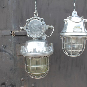 May include: Two silver industrial cage lights hanging from chains against a dark gray metal background. The lights have a clear glass globe and a wire cage around the bulb. The lights are both turned off.