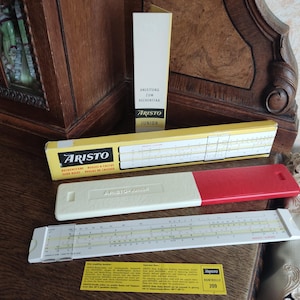 May include: A vintage slide rule with a white body and red accents. The slide rule is labelled "ARISTO JUNIOR" and is packaged in a yellow box with the brand name "ARISTO".