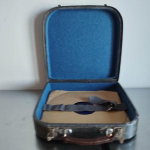 Könnte beinhalten: Geöffnete, vintage Schallplattenkoffer mit blauem Innenraum und braunem Griff. Der Koffer ist grau mit silberfarbenen Metallakzenten. Im Inneren ist eine Schallplatte mit einem grauen Riemen gesichert.