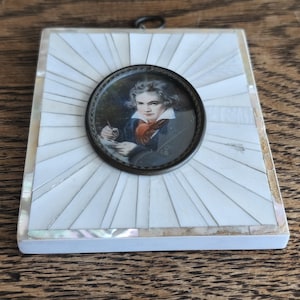 Puede incluir: Miniatura de retrato antiguo, de forma cuadrada, con un retrato de un hombre con abrigo oscuro y bufanda roja. El retrato redondo está centrado en un fondo cuadrado de baldosas de nácar blancas. Un pequeño anillo de metal en la parte superior.