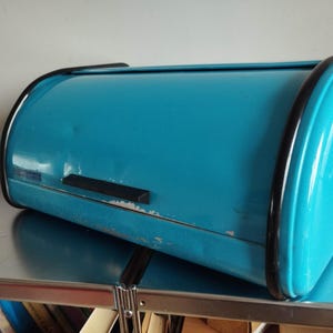 Könnte beinhalten: Ein leuchtend türkisfarbener Brotkasten aus Metall mit Rolldeckel, schwarzer Umrandung an den Kanten und einem schwarzen Griff. Die Außenfläche weist leichte Lackabnutzungen und kleine Dellen auf und hat eine glänzende Oberfläche.