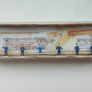 May include: A set of six miniature figures of train station personnel, including a conductor, a station master, and a ticket collector. The figures are painted in blue and white uniforms and are standing on a platform in front of a train.