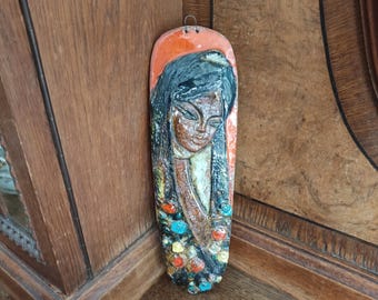 May include: A ceramic wall hanging with a relief sculpture of a woman's face. The woman has long black hair and is wearing a colorful patterned garment. The background is a light orange color.