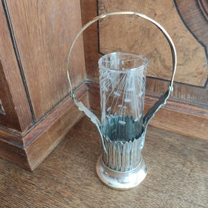 Puede incluir: Jarrón de flores plateado con un inserto de vidrio. El jarrón tiene un asa y un diseño decorativo. El inserto de vidrio tiene un patrón floral.