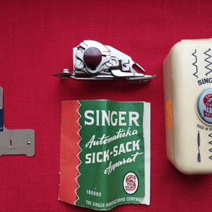 Könnte beinhalten: Eine Sammlung von Vintage-Zubehör für Singer-Nähmaschinen. Enthält einen Metall-Nähfuß, ein cremefarbenes Gehäuse mit dem Singer-Logo und ein grün-rotes Anleitungsheft. Die Artikel befinden sich auf rotem Hintergrund.