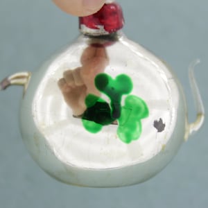 Puede incluir: Un adorno de vidrio plateado con forma de tetera con un diseño de trébol verde.