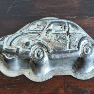 Puede incluir: Molde de metal vintage con forma de coche, con un acabado plateado desgastado. El molde presenta contornos detallados del coche, incluyendo ventanas y ruedas. El molde para coche es probablemente para hornear o manualidades.