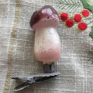 Puede incluir: Un adorno de Navidad de vidrio vintage con forma de hongo, con un sombrero marrón y un tallo blanco. El adorno tiene un clip de metal para colgar.