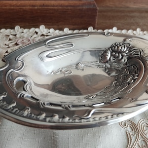 Puede incluir: Un plato ovalado de plata con un diseño ornamentado que presenta el rostro de una mujer y motivos florales. El plato tiene un borde elevado y una superficie lisa y pulida.