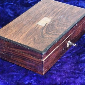 Può includere: Una scatola rettangolare in legno con finitura marrone scuro e una piccola placca metallica rettangolare sul coperchio. La scatola ha una serratura e una piccola chiave. La scatola viene probabilmente utilizzata per la conservazione.