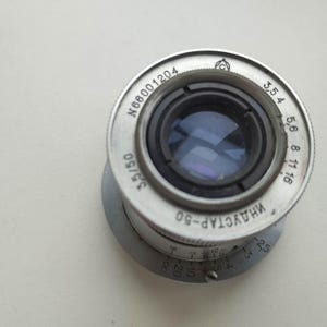 Puede incluir: Un objetivo de c&aacute;mara Industar-50 de color plateado con componentes internos negros. El objetivo tiene una serie de n&uacute;meros y marcas alrededor del borde, incluyendo "3,5/50" y "N66001204". El objetivo est&aacute; sobre un fondo blanco.