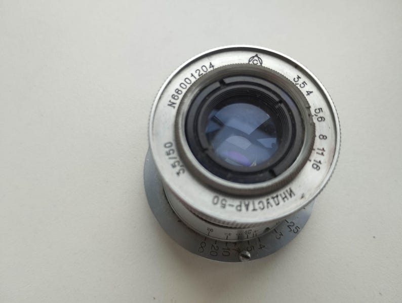 Puede incluir: Un objetivo de c&aacute;mara Industar-50 de color plateado con componentes internos negros. El objetivo tiene una serie de n&uacute;meros y marcas alrededor del borde, incluyendo "3,5/50" y "N66001204". El objetivo est&aacute; sobre un fondo blanco.