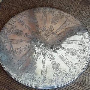 Puede incluir: Una bandeja circular de plata con un intrincado diseño floral. La bandeja tiene un centro elevado y un borde festoneado.