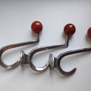 Può includere: Tre ganci da parete in metallo argentato lucido, ciascuno caratterizzato da un design a gancio curvo e da un terminale a sfera rosso scuro lucido sulla parte superiore. I ganci presentano una piastra di montaggio circolare con fori.