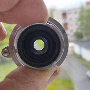 Puede incluir: Primer plano de un componente de lente de metal plateado con un interior negro y una abertura central. La lente est&aacute; sujeta por una mano, con un fondo borroso al aire libre visible.