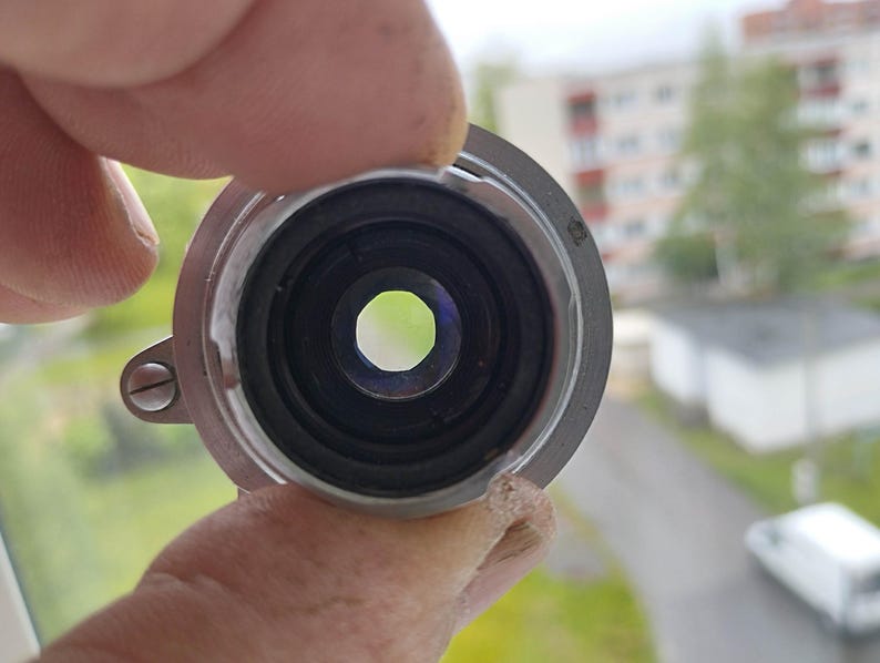 Puede incluir: Primer plano de un componente de lente de metal plateado con un interior negro y una abertura central. La lente est&aacute; sujeta por una mano, con un fondo borroso al aire libre visible.