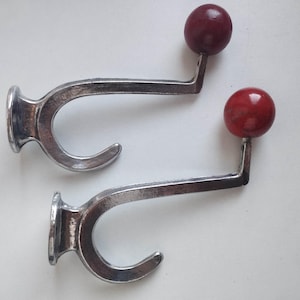 Pode incluir: Dois ganchos de parede feitos de metal prateado polido, cada um com um gancho inferior curvo e um braço reto que se estende para cima até um remate esférico liso de cor vermelho-escura. As superfícies metálicas têm um acabamento reflexivo.