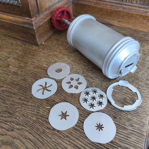 Puede incluir: Una prensa de galletas plateada vintage con un mango rojo y varias formas de disco decorativas. La prensa es cilíndrica e incluye varios discos intercambiables para crear diferentes diseños de galletas. Los discos son redondos con patrones de estrella, flor y otros.