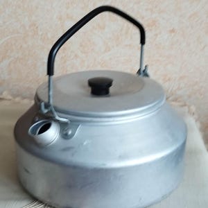 May include: A silver aluminum kettle with a black handle and a lid. The kettle has a spout on the side and a small black knob on the lid.