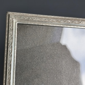 May include: A rectangular picture frame with a metallic silver finish and a repeating embossed geometric pattern along the border. The frame contains a clear glass pane covering a dark grey textured fabric matting with a visible light reflection.