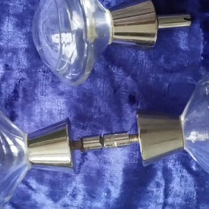 Puede incluir: Dos pomos de puerta de cristal transparente con un diseño acanalado y bases de metal plateado. Los pomos están colocados sobre un fondo azul texturizado.