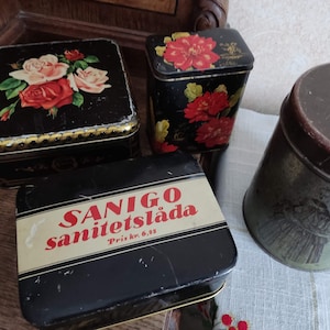 以下が含まれることがあります： 花柄とテキストのデザインが施されたヴィンテージの金属製の缶4つ。缶は黒で金色の縁取りがされており、赤と白の花が描かれています。1つの缶には「SANIGO Sanitetsläda Pris kr. 6.35」というテキストが書かれています。