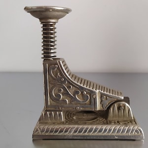 May include: A silver metal paperweight with an ornate design and a spring-loaded mechanism. The paperweight has a decorative base and a top with a small, round opening.