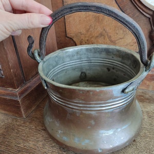 Può includere: Una pentola di rame vintage con un manico in pelle marrone scuro usurato. La pentola ha un corpo arrotondato e una parte superiore larga e aperta. Il manico è attaccato alla pentola con due anelli di metallo.
