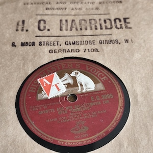 May include: Vintage record label with a red and black design, featuring a white dog with a gramophone. The label is from the "His Master's Voice" series, with text in German. The record is housed in a tan paper sleeve.