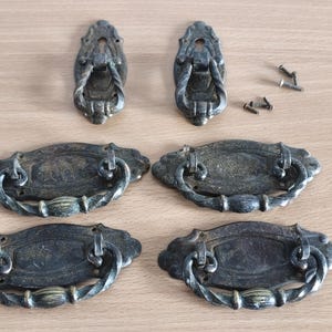 Op de afbeelding: Antieke bronzen lade- en handgrepen. De set bevat vier ovale handgrepen met gebogen handgrepen en twee kleinere, sierlijke handgrepen. Er zijn ook verschillende schroeven zichtbaar.