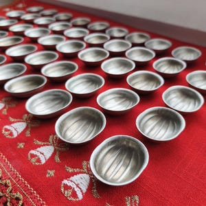 May include: A collection of small, silver-colored metal bowls arranged on a red fabric with a decorative pattern. The bowls have a ribbed interior design and are evenly spaced across the surface.