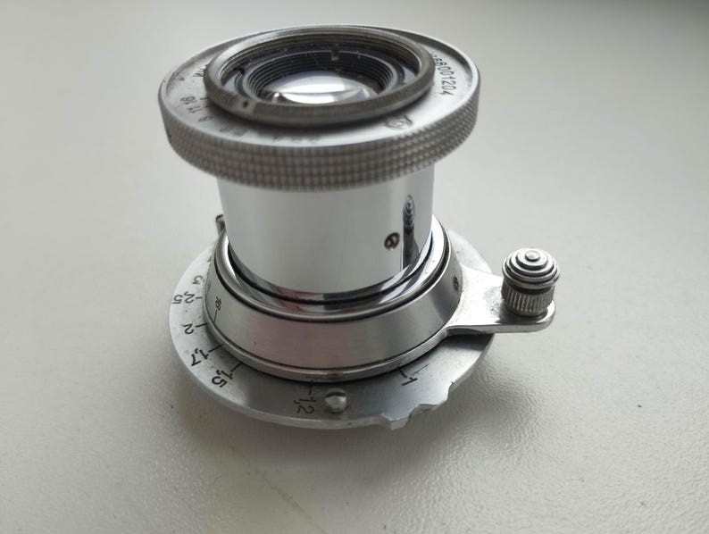 Puede incluir: Un objetivo de c&aacute;mara plateado vintage con un anillo de enfoque moleteado y ajustes de apertura. El objetivo tiene una superficie brillante y reflectante y una serie de n&uacute;meros y marcas. El objetivo est&aacute; montado sobre una base circular con marcas adicionales y un peque&ntilde;o bot&oacute;n de ajuste.