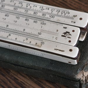 May include: Close-up of a vintage slide rule with white scales and black markings. The slide rule is partially resting on a worn, dark green case. The text "COPYRIGHT" is visible.