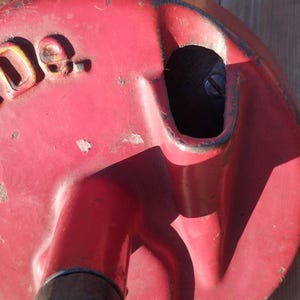 May include: Red metal object with a handle and a circular top. The top has a raised inscription that reads "PE-DE."