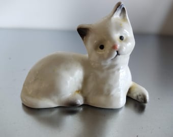 May include: White ceramic figurine of a cat lying down with its head turned to the side. The cat has black ears and a pink nose.