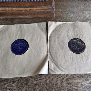 May include: Two vintage Columbia records in paper sleeves. The record labels are dark blue with white text. The sleeves are a light tan color. The records are on a wooden surface.