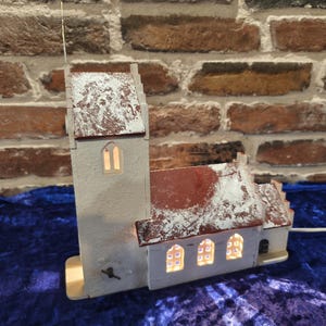 Peut inclure: Une maquette d'église miniature avec une haute tour et une section de bâtiment plus petite. Le modèle est peint en gris clair avec des accents rouges sur le toit, et les fenêtres sont éclairées. L'église est posée sur une surface bleue.