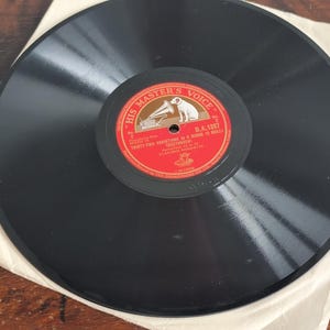 May include: A vintage black vinyl record with a red and gold label featuring the text "HIS MASTER'S VOICE" and a dog listening to a gramophone. The record is in a paper sleeve, resting on a wooden surface.