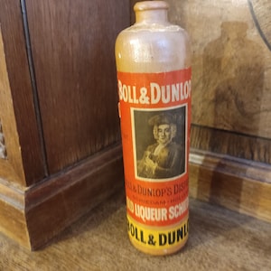 Può includere: Una bottiglia vintage in ceramica marrone con un'etichetta che recita "Boll & Dunlop's Distilled Old Liqueur Schnapps Boll & Dunlop". L'etichetta presenta un ritratto di un uomo con una parrucca in polvere e un'immagine in bianco e nero di una bottiglia.
