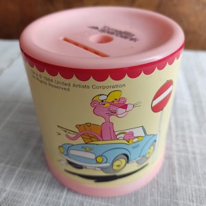 Könnte beinhalten: Rosa Plastik-Sparschwein mit dem Pink Panther, der ein blaues Auto fährt. Das Sparschwein hat ein rot-weiß gestreiftes Design mit dem Text "TM & C 1984 United Artists Corporation All Rights Reserved".